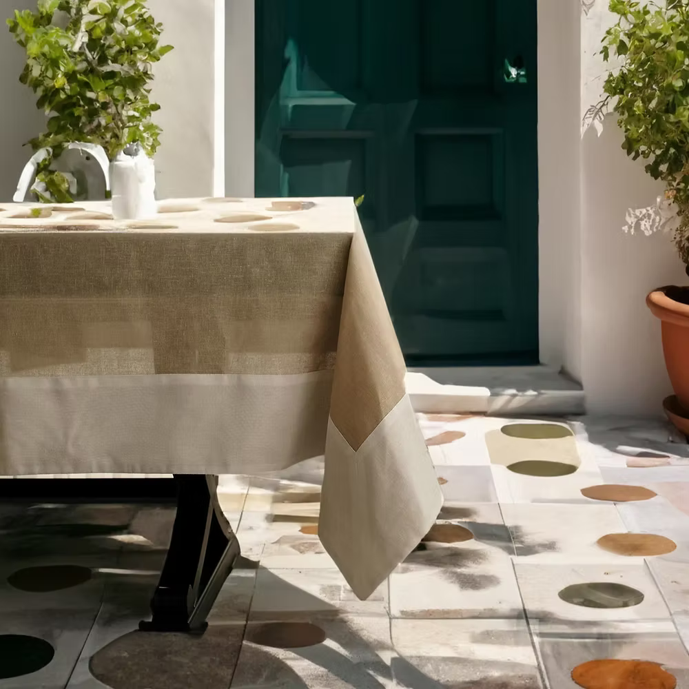 22 Maggio Istanbul Terreno Linen Tablecloth-Table with a beige tablecloth on a tiled floor with decorative patterns, surrounded by plants.