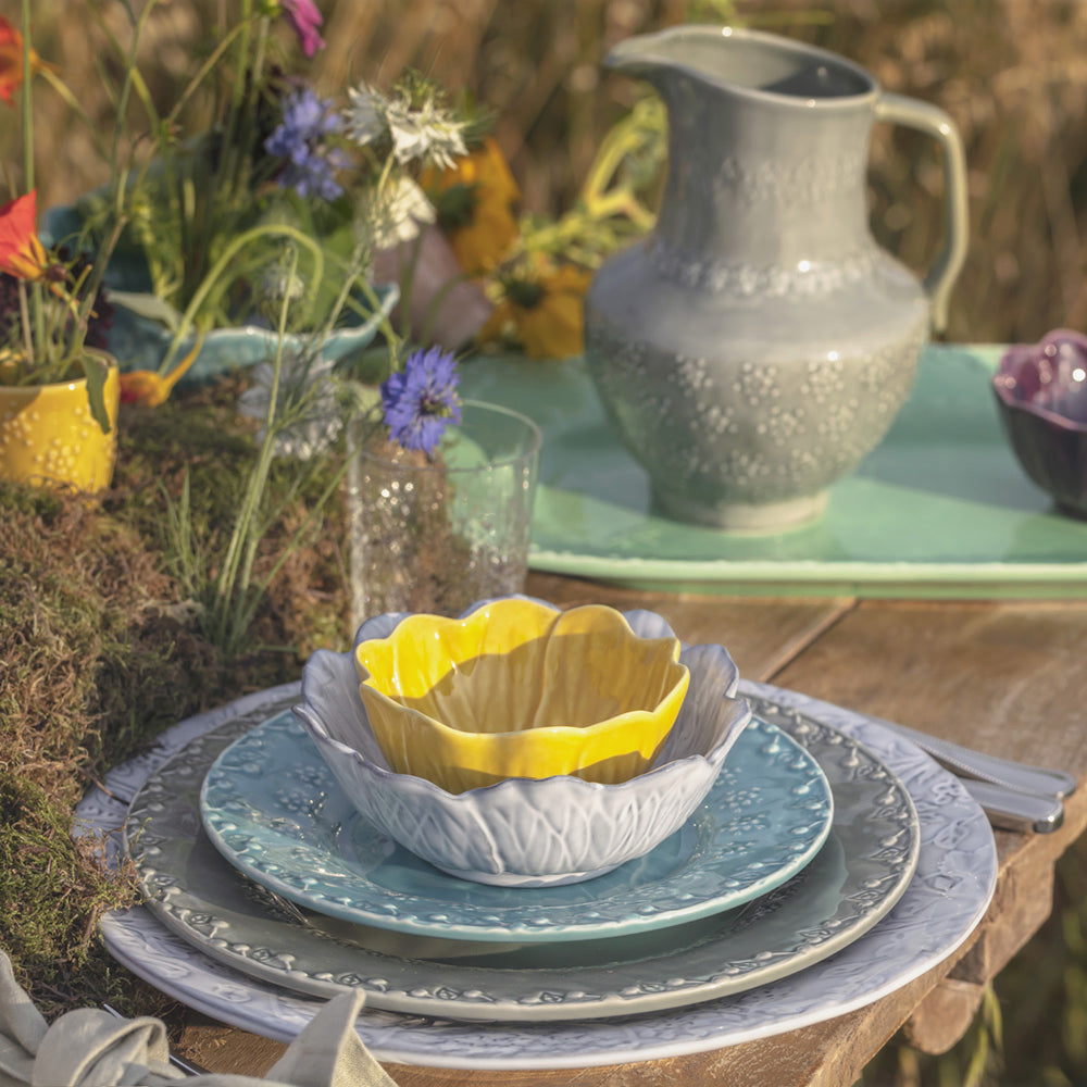 Bordallo Pinheiro Flora Dessert Plate-Decorative table setting with a yellow bowl, blue plates, and a gray pitcher on a wooden surface.