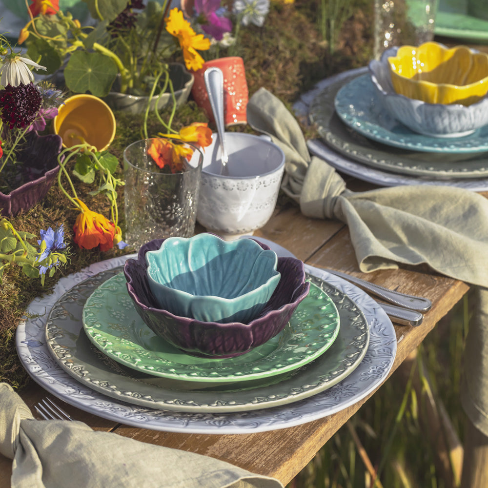 Bordallo Pinheiro Flora Dessert Plate-Table setting with colorful ceramic bowls and plates on a wooden table, surrounded by flowers.