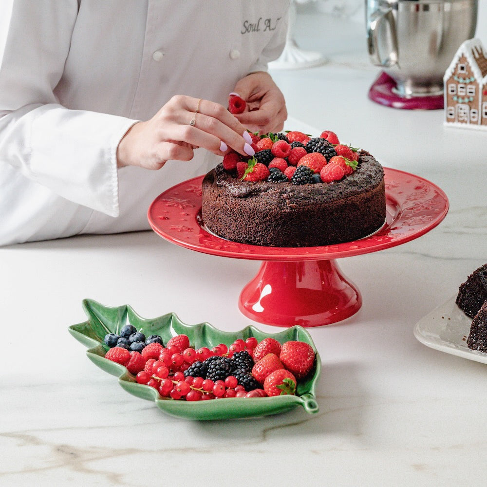 Bordallo Pinheiro Holly Leaf Platter-Person decorating a chocolate cake with berries on a red cake stand, with a green berry plate nearby.