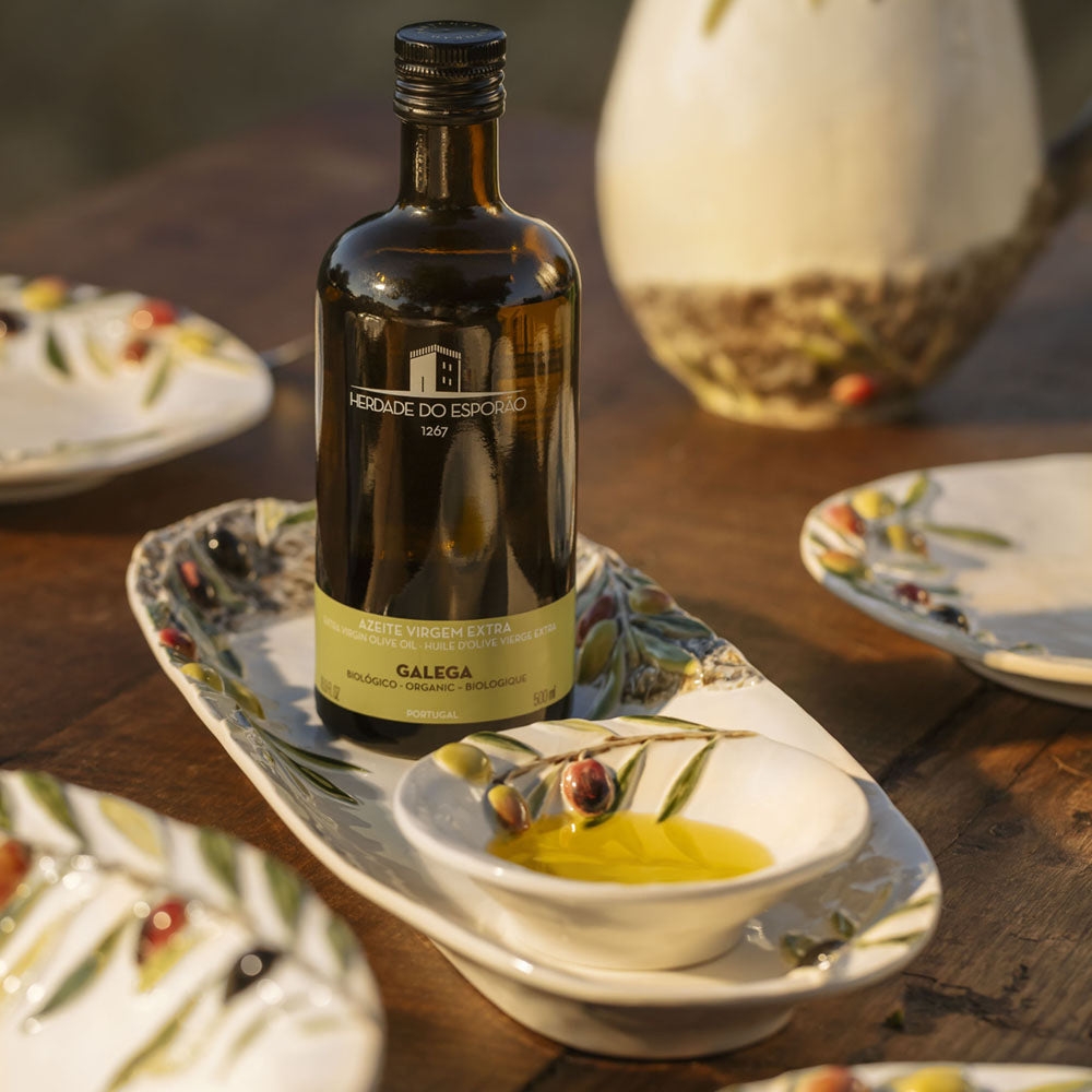 Bordallo Pinheiro Olival Platter-Bottle of olive oil on a wooden table with a bowl of olive oil and plates in the background.