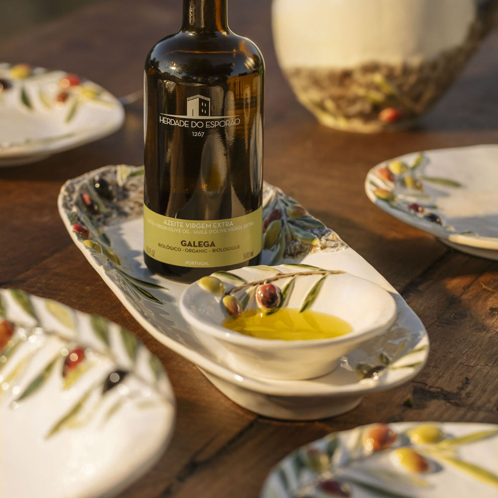 Bordallo Pinheiro Olival Small Bowl-Bottle of olive oil on a decorative plate with olive oil, surrounded by plates with olive designs on a wooden table.