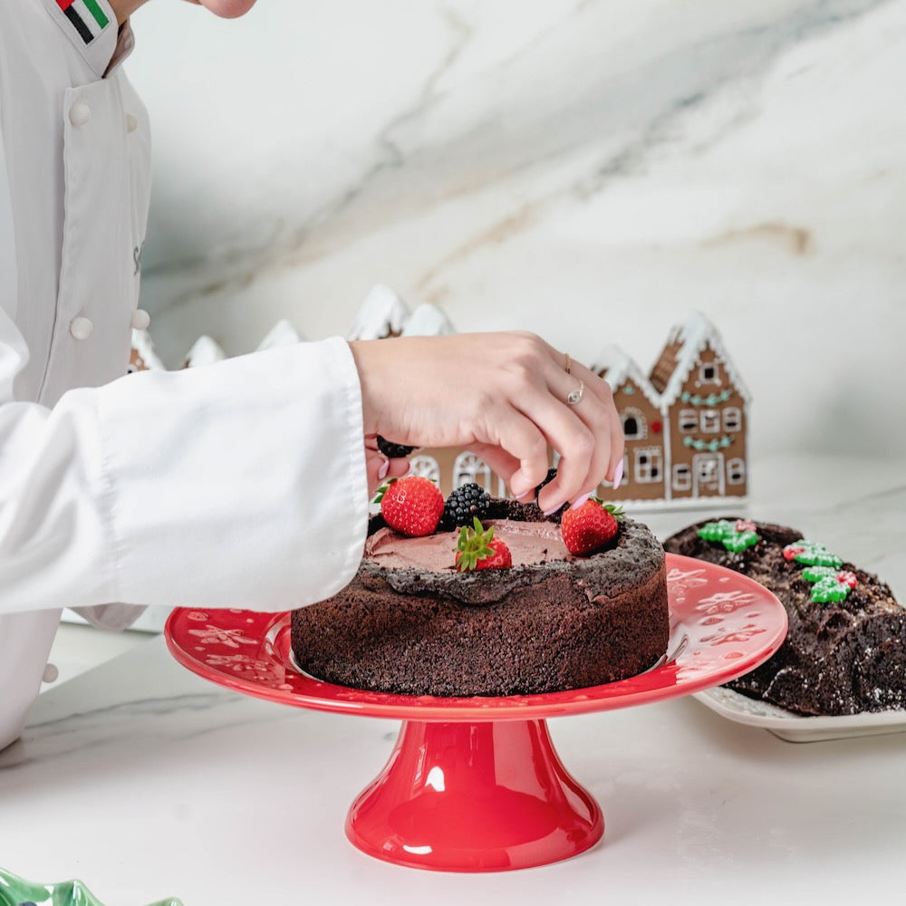 Bordallo Pinheiro Snowflakes Cake Stand-Person decorating a chocolate cake with berries on a red cake stand, with a gingerbread house in the background.