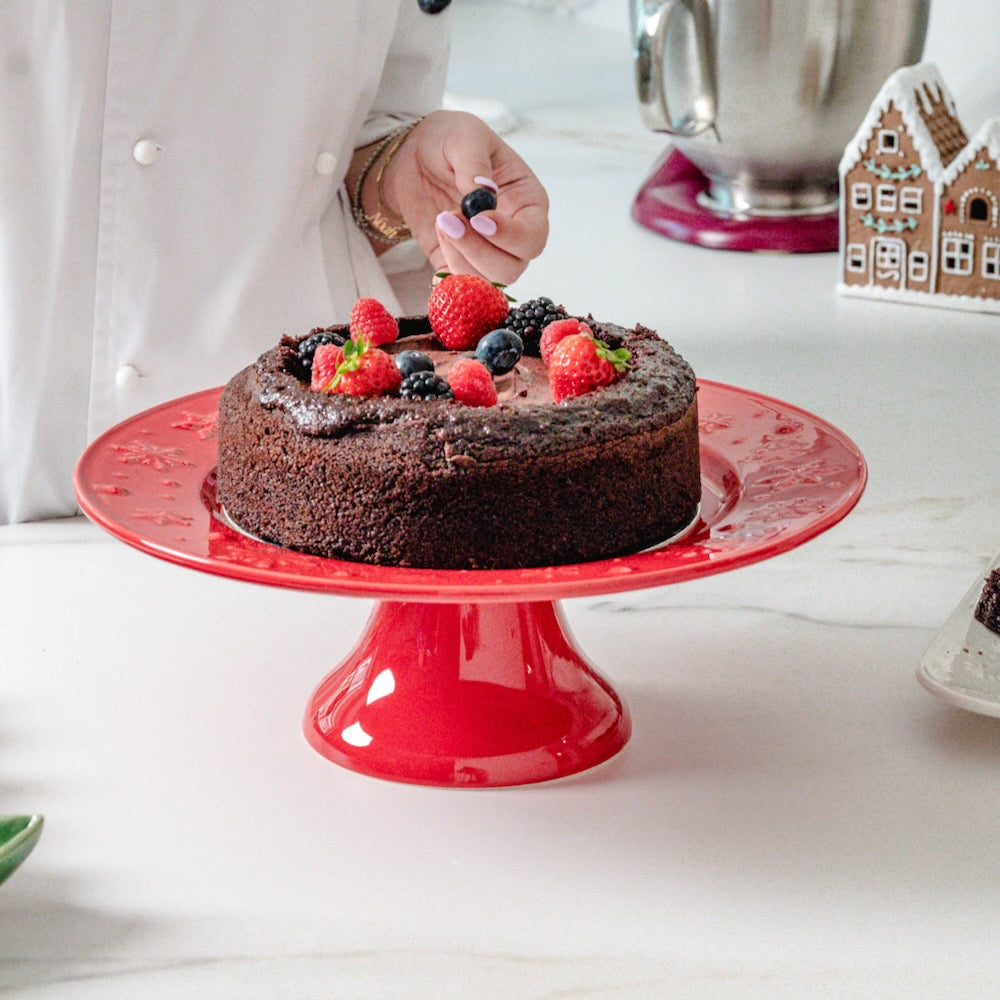 Bordallo Pinheiro Snowflakes Cake Stand-Chocolate cake with berries on a red cake stand, being decorated by a person.