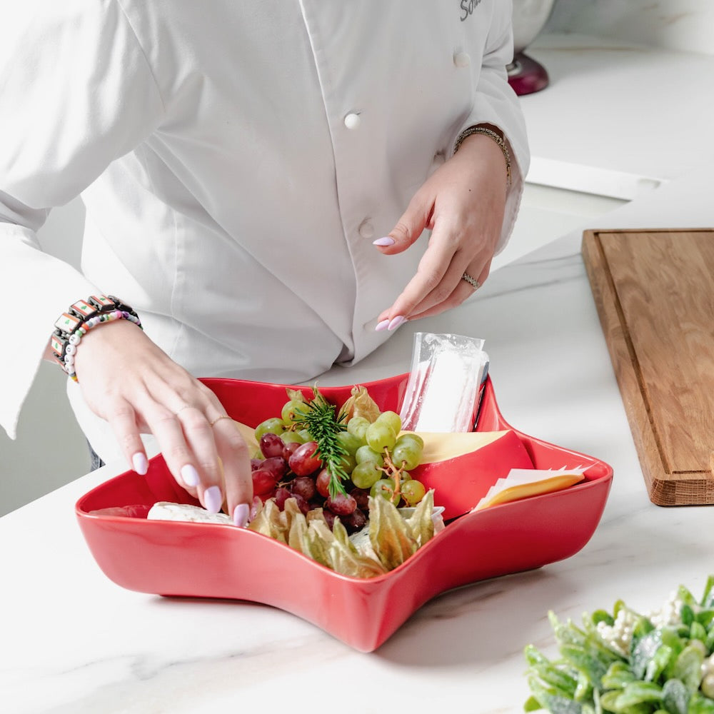Bordallo Pinheiro Snowflakes Star Bowl, 37cm-Person arranging fruits and cheese on a red star-shaped platter in a kitchen setting.