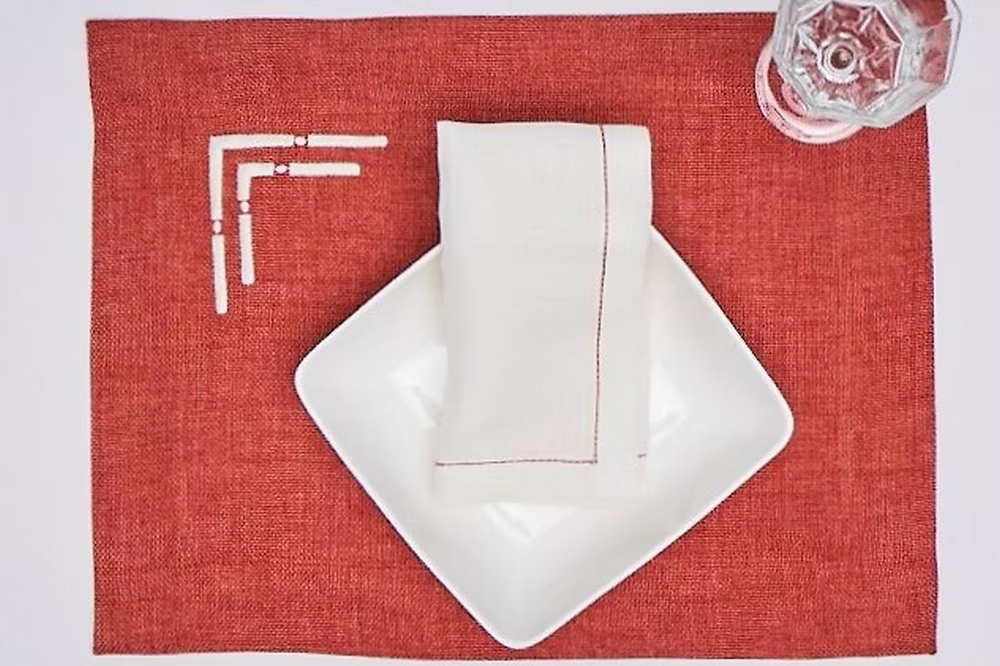 Doppia Linen Napkin Set of 4-White square plate with folded white napkin on a red placemat, with a glass of water in the corner.