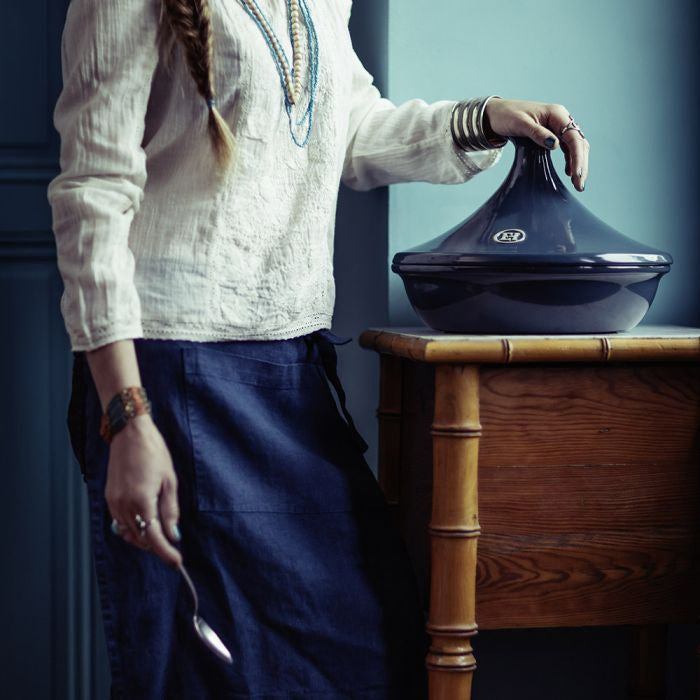 Emile Henry Flame Tagine-Person holding a dark blue pot on a wooden table against a blue wall.