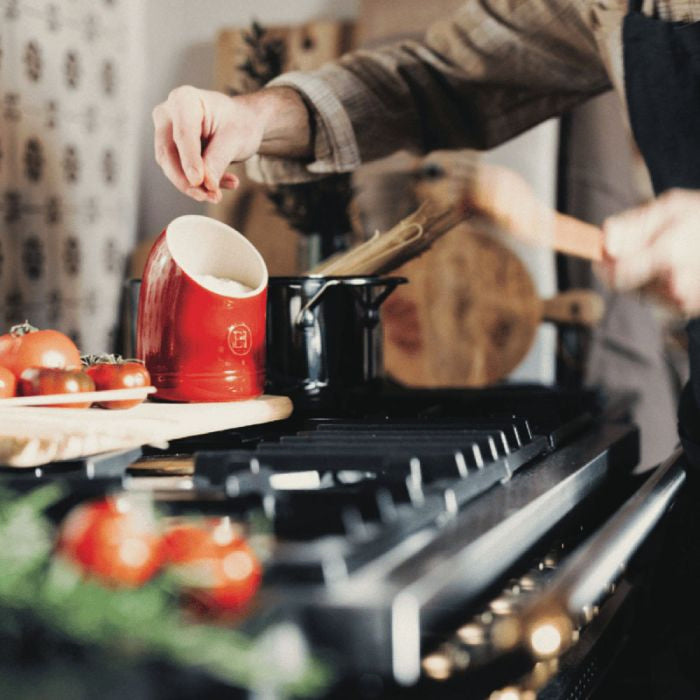 Emile Henry Salt Keeper-Person cooking on a stove with a red cup and tomatoes on a wooden board.