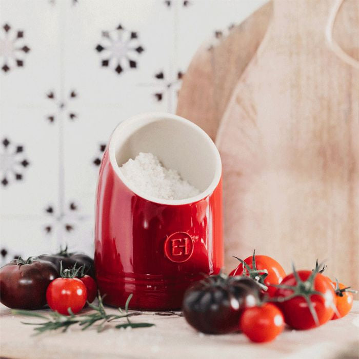 Emile Henry Salt Keeper-Red pepper grinder with white top on a wooden surface with tomatoes and herbs