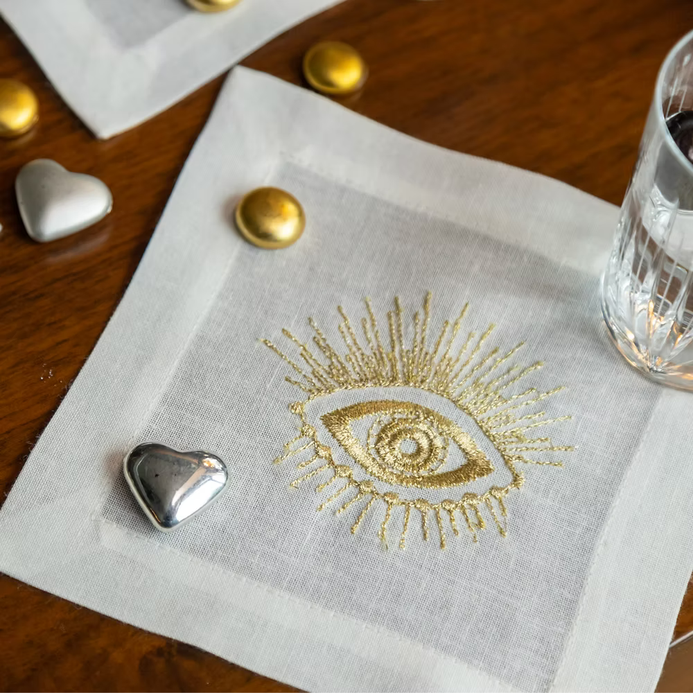 Fatima Linen Coasters Set of 4-White napkin with gold embroidered eye design on a wooden table.