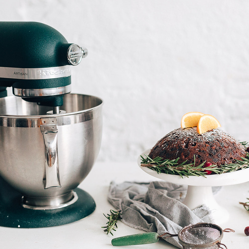 KitchenAid Green stand mixer with a silver bowl next to a decorated Christmas pudding on a white surface.