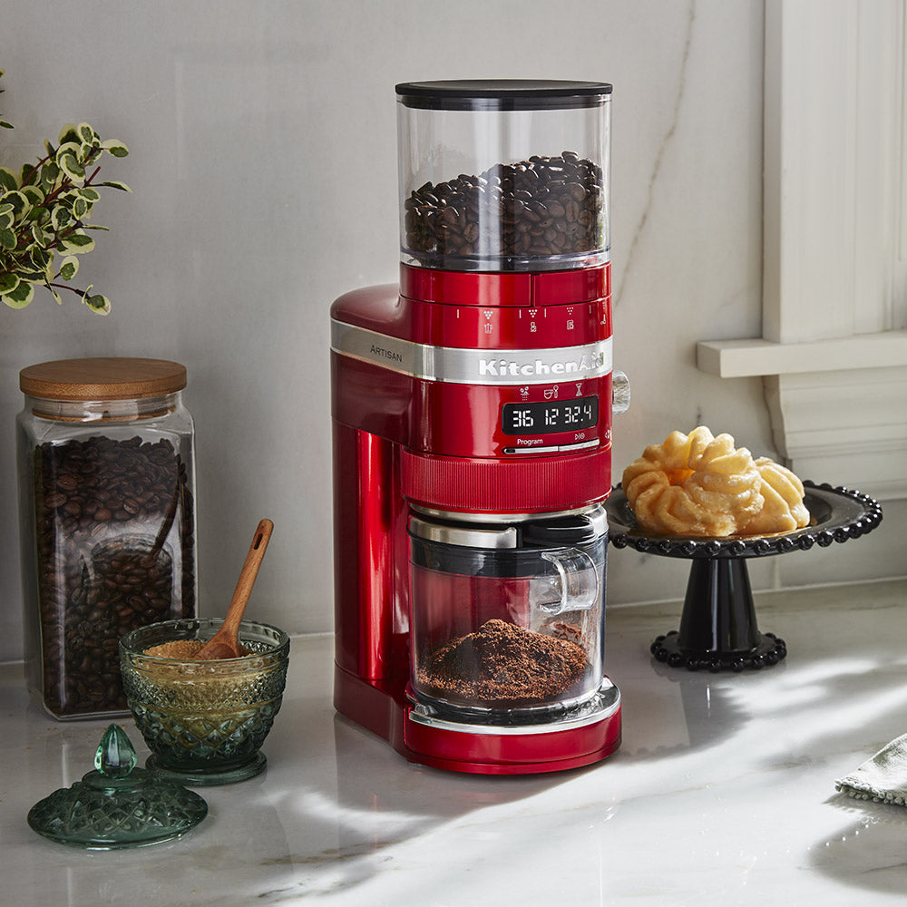 Red coffee grinder on a kitchen counter with coffee beans and a jar of ground coffee.