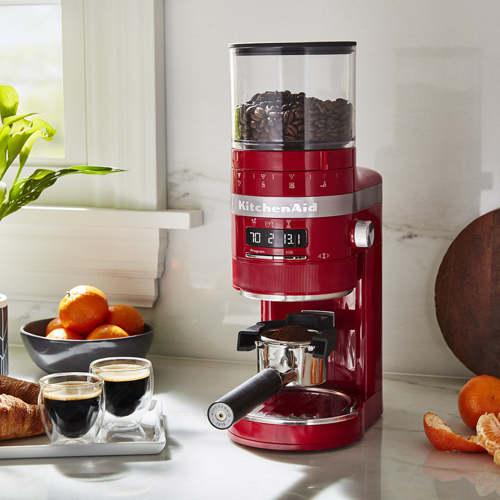 Red KitchenAid coffee grinder on a kitchen counter with coffee and oranges.