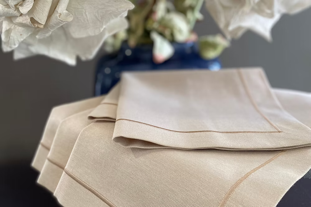 Latte Dinner Napkin Set of 4-Beige folded fabric with floral arrangement in the background