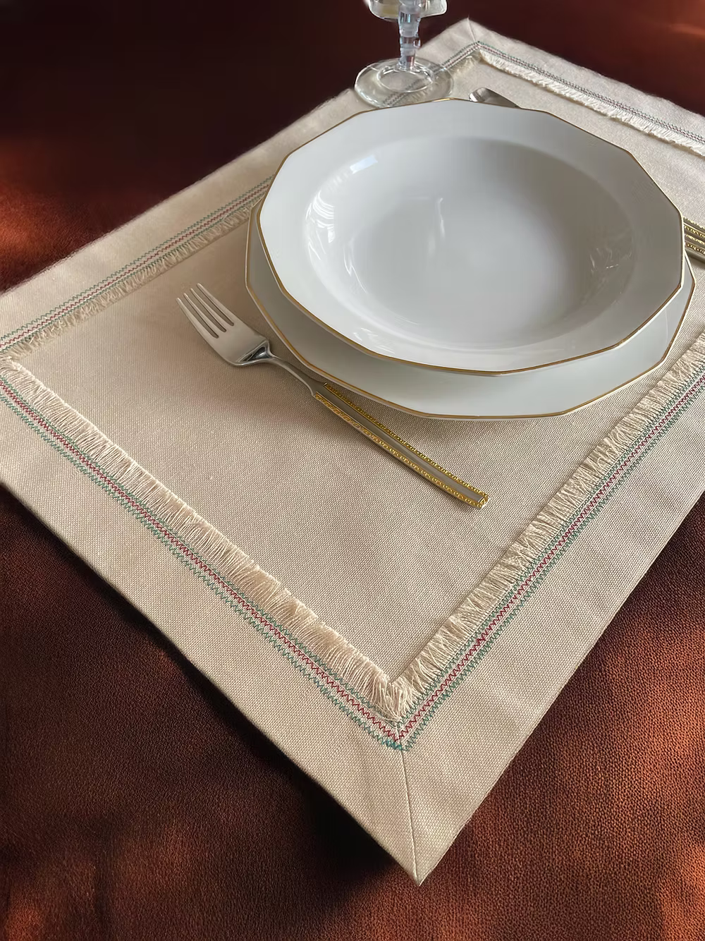 Nappina Placemat Set of 4-White ceramic plates with gold rims on a beige placemat with decorative edge, on a wooden table.