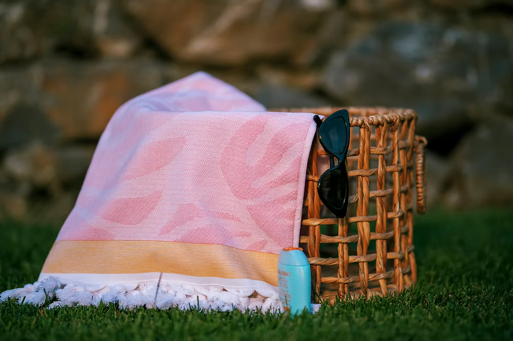 Premium Vivace Cotton Beach Towel-Towel, sunglasses, and sunscreen on a grassy area with a stone wall background