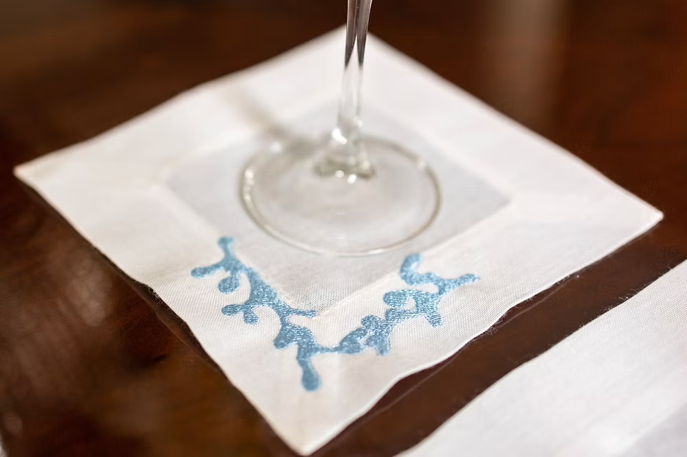 Sirene Linen Coasters Set of 4-Clear glass on a white napkin with blue designs on a wooden surface