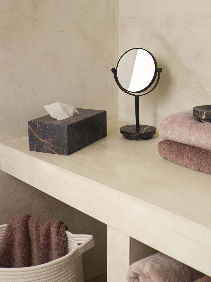Small round mirror on a stand next to a tissue box and folded towels on a light-colored surface.
