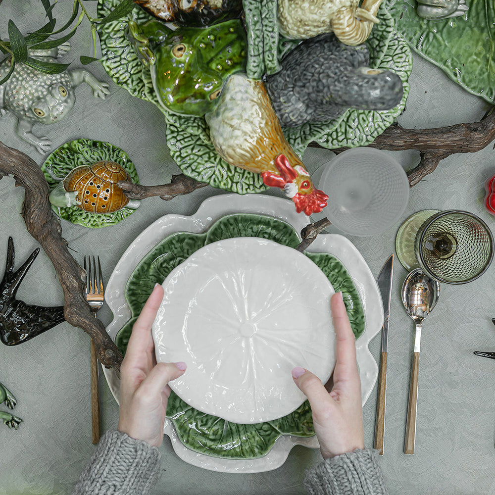 Person holding a white plate over a decorative green plate with animal designs on a table.