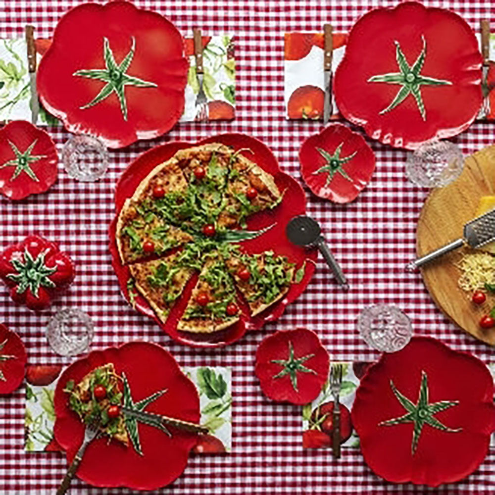 Table setting with pizza on a red plate, surrounded by tomato decorations on a red and white checkered tablecloth.