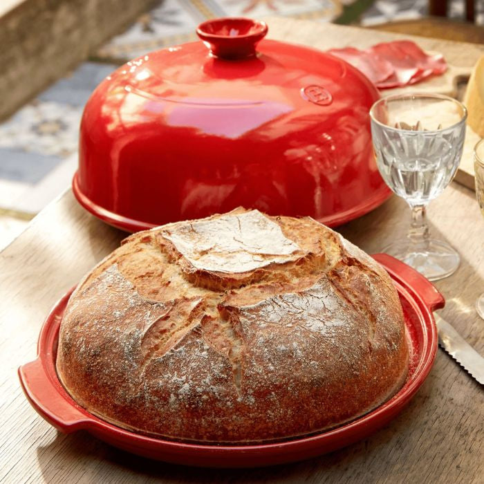 Emile Henry Bread Baker-Loaf of bread in a red ceramic dish on a wooden table with a glass of water.