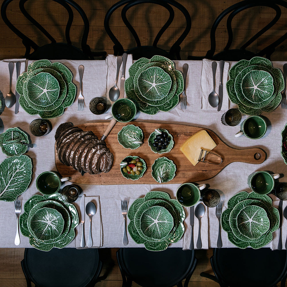 Dining table set with green ceramic plates, a wooden cutting board with food, and silverware on a white tablecloth.