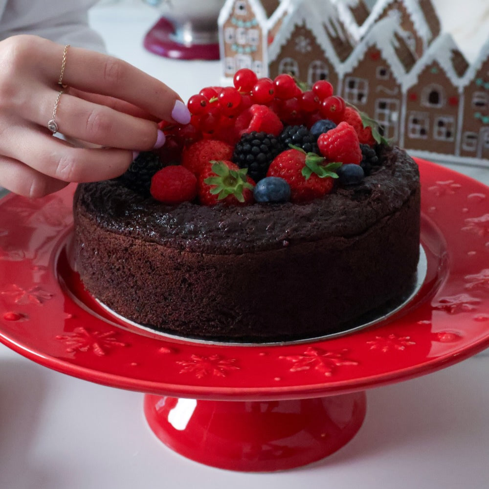 Chocolate cake with berries on a red cake stand, with a gingerbread house in the background.
