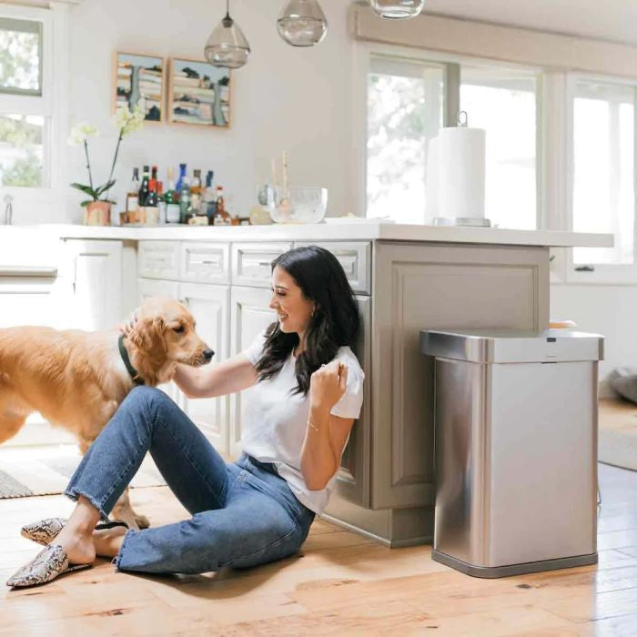 simplehuman Voice and Motion Sensor Bin, Rectangular-Woman sitting on the floor with a dog in a kitchen next to a stainless steel trash can.