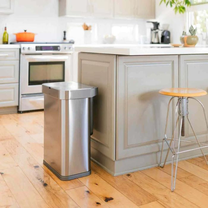 simplehuman Voice and Motion Sensor Bin, Rectangular-Stainless steel trash can in a kitchen setting with wooden floor and gray cabinets.