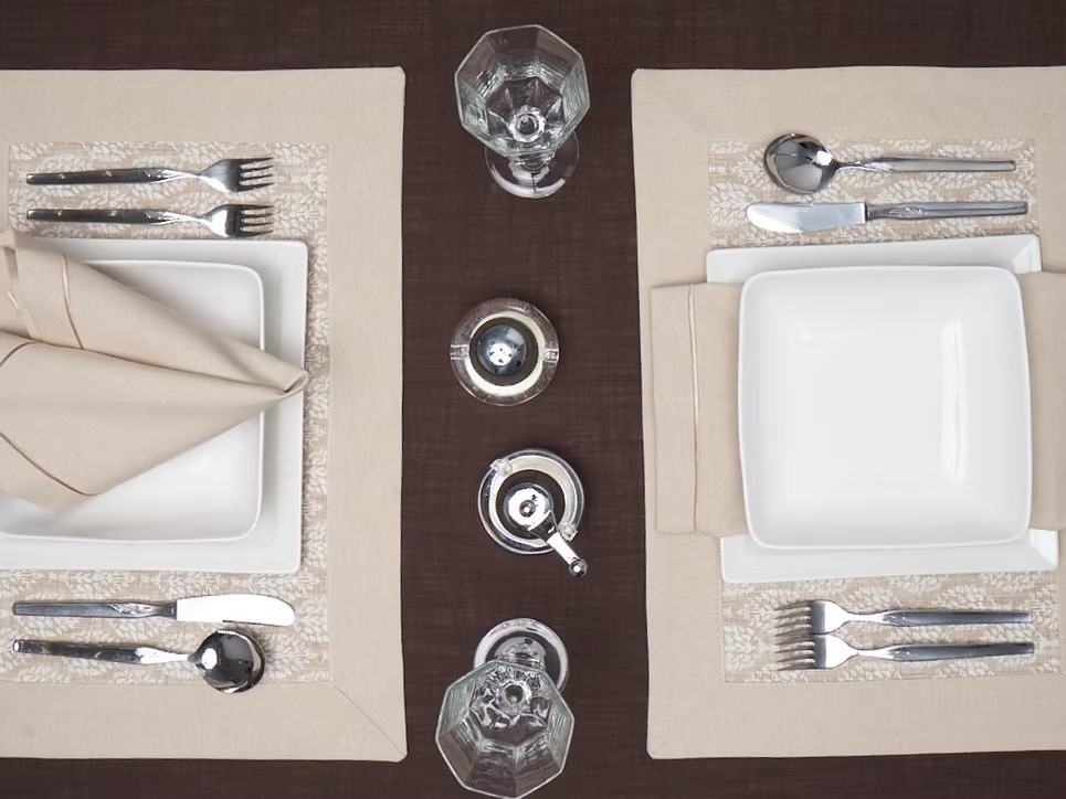 22 Maggio Istanbul Latte Placemat Set of 4-Elegant table setting with white plates, silverware, and glasses on a brown tablecloth.
