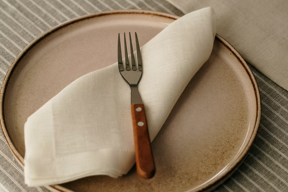 22 Maggio Istanbul Terreno Beige Linen Napkin Set of 4-Beige ceramic plate with a folded white napkin and a fork on a textured surface