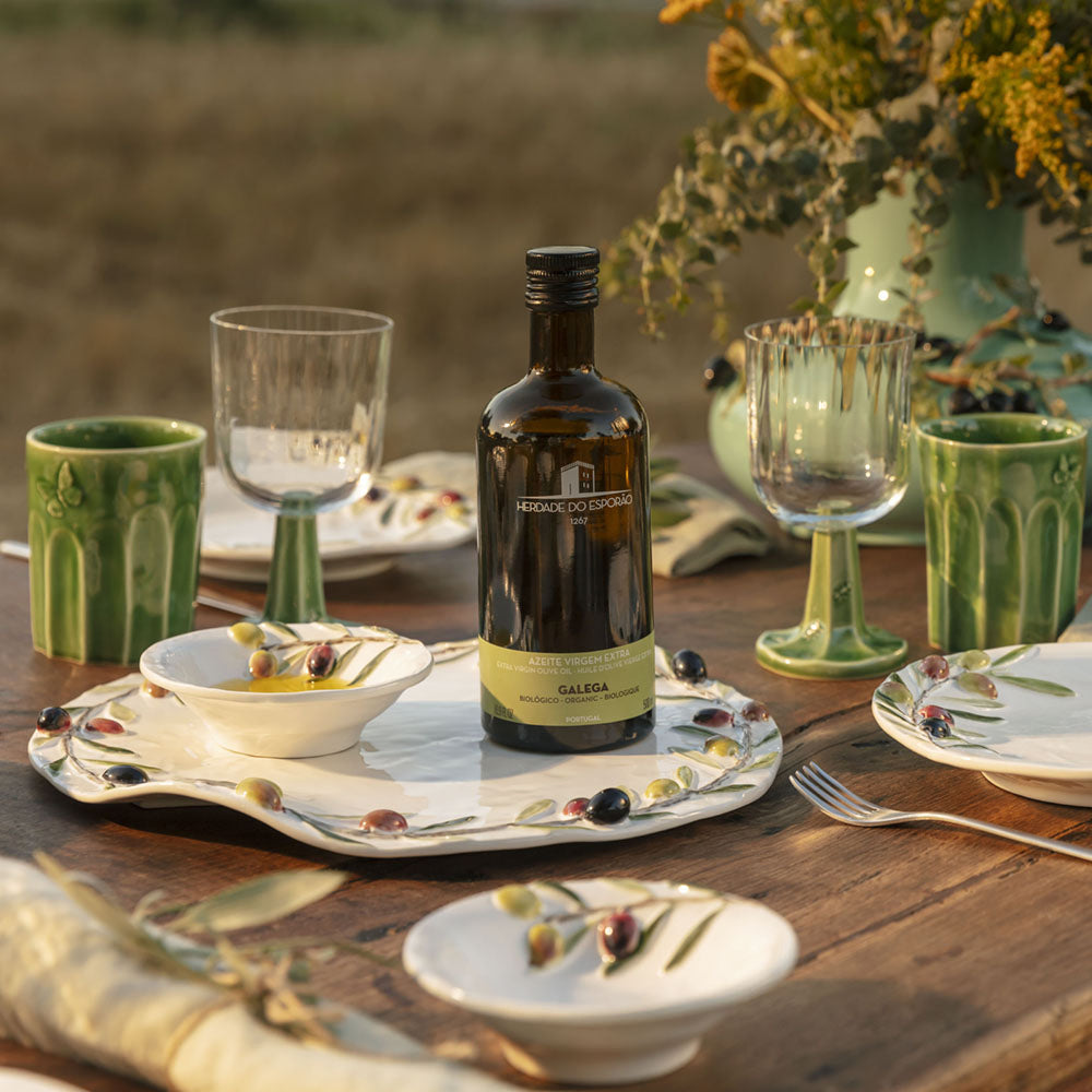 Bordallo Pinheiro Olival Cheese Platter-Table setting with a bottle of olive oil, glasses, and plates on a wooden table outdoors.