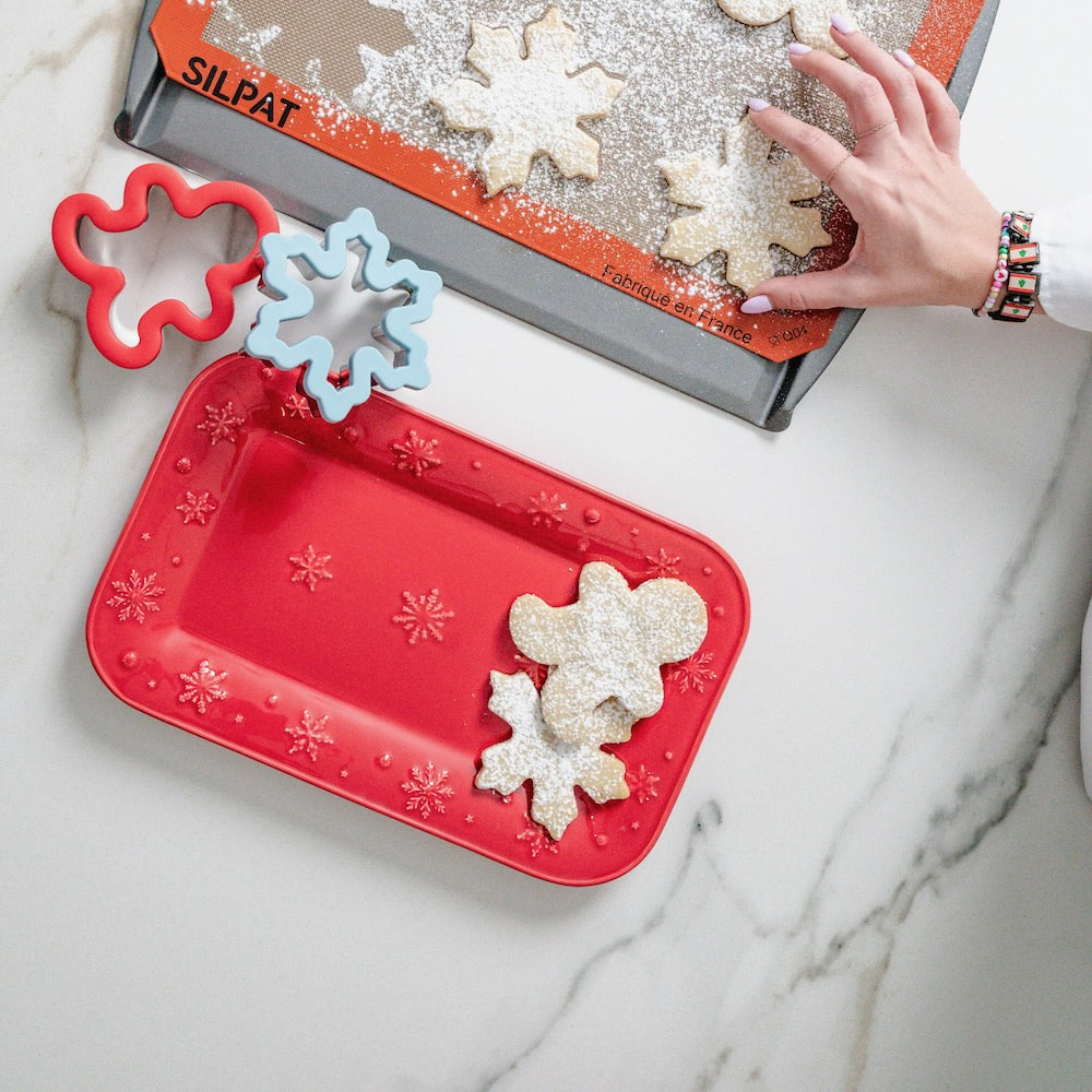 Bordallo Pinheiro Snowflakes Platter-Decorative red cookie sheet with snowflake designs next to a baking tray with cookies and cookie cutters.