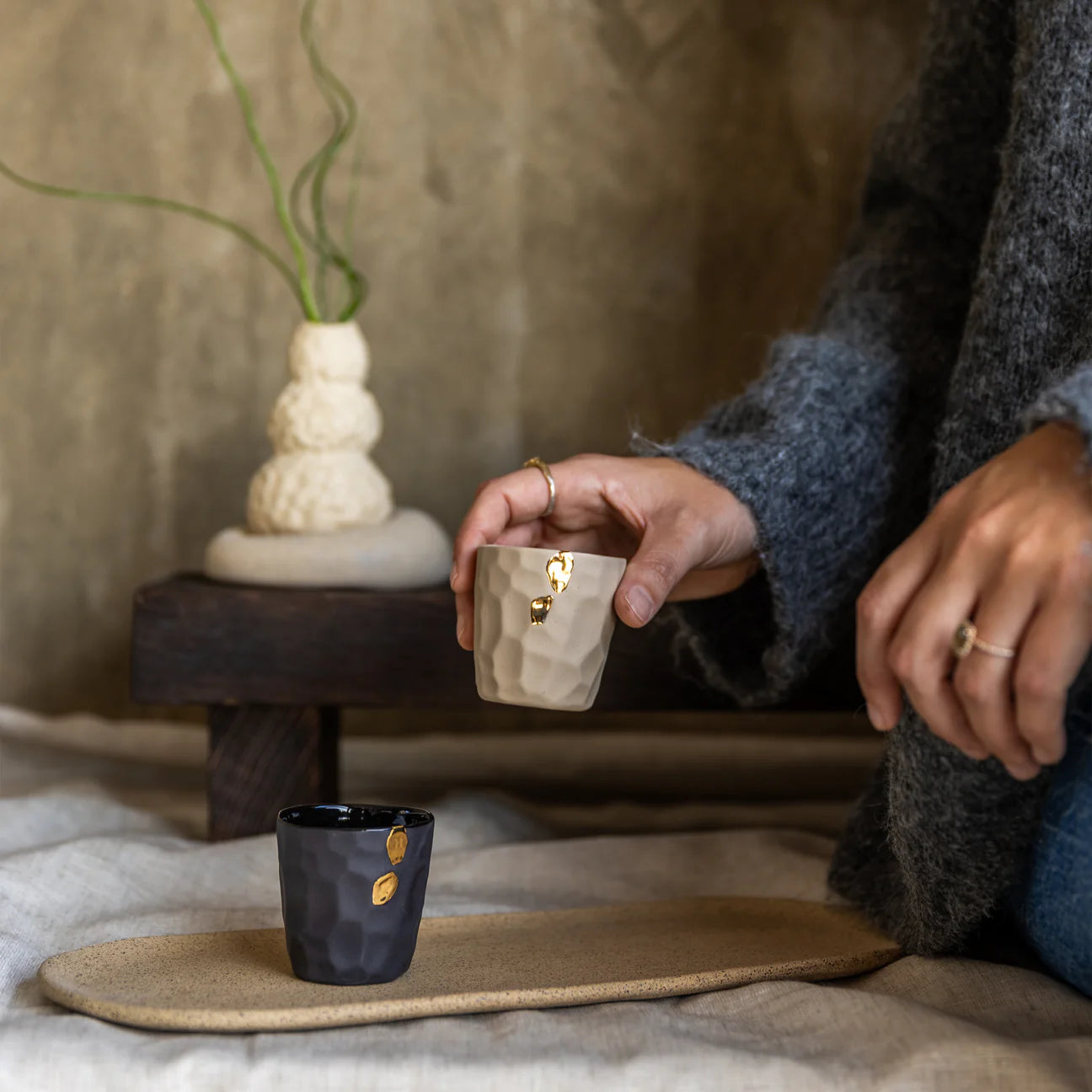 Person holding a small white ceramic Cawa Cups with gold accents on a textured surface.
