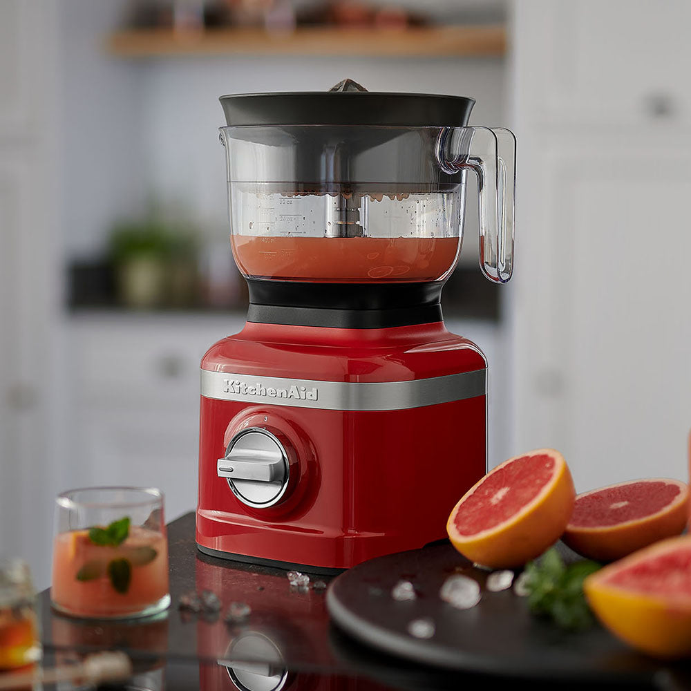 Red KitchenAid blender with a glass container filled with a red liquid, surrounded by sliced grapefruits on a kitchen counter.