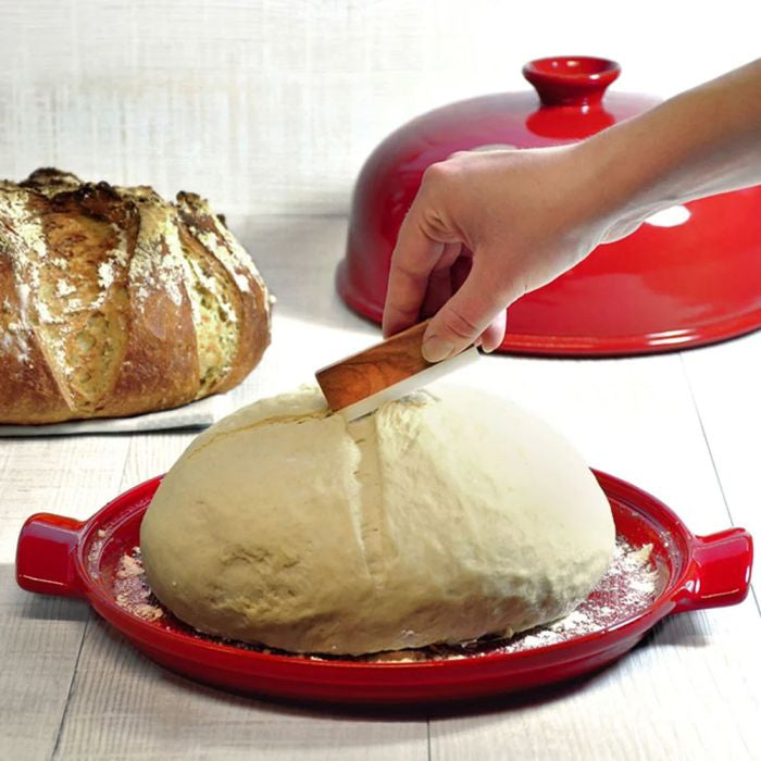 Emile Henry Baker's Blade-Person cutting a block of cheese with a knife on a red cheese slicer, with a loaf of bread in the background.