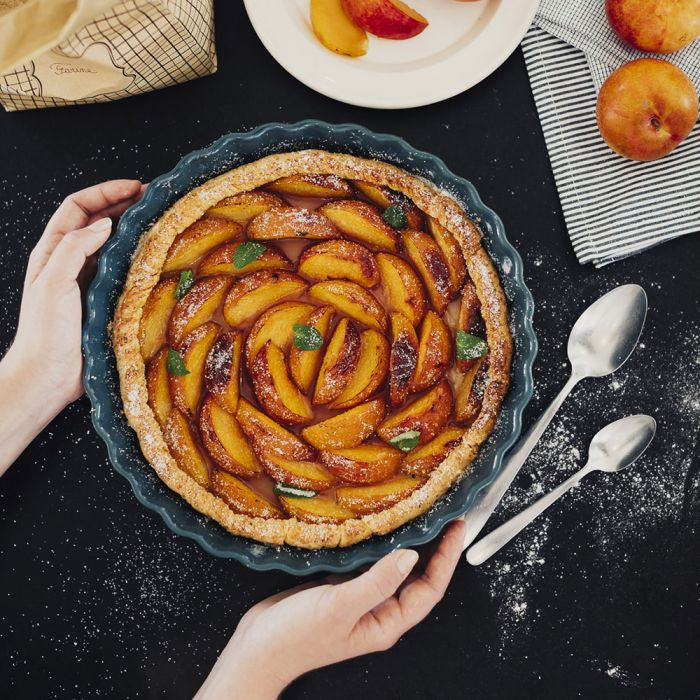 Peach tart held by hands on a dark surface with utensils and peaches.