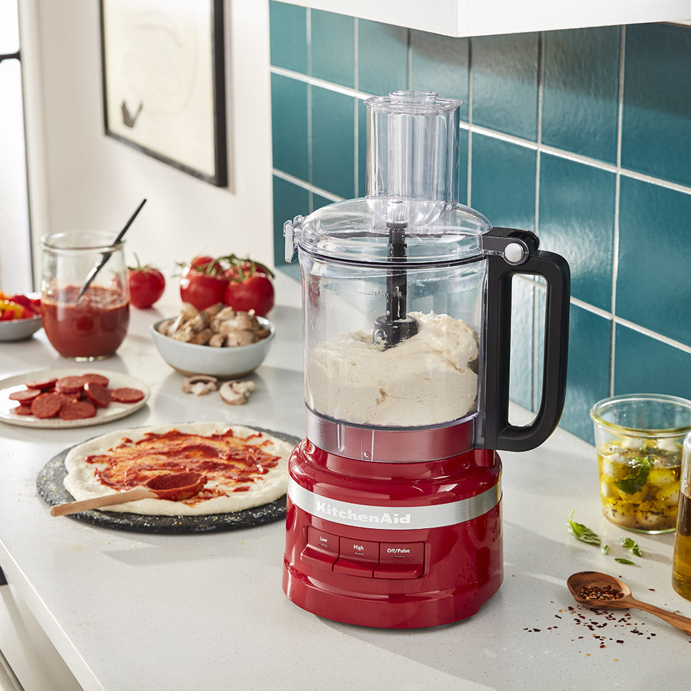 Red KitchenAid food processor on a kitchen counter with ingredients and a pizza in the background.