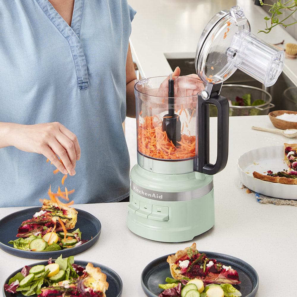 Person preparing a salad with a KitchenAid food processor on a kitchen counter.