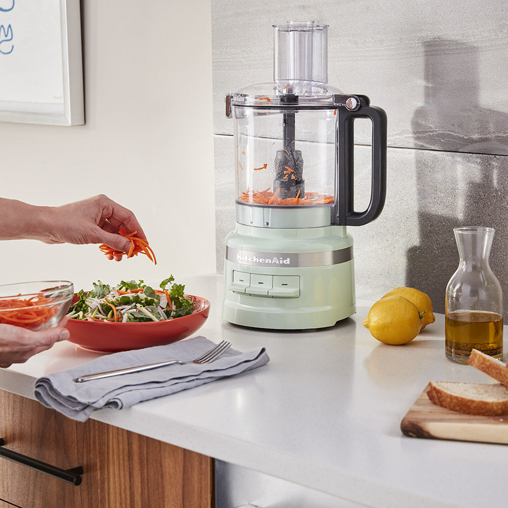 Kitchenaid food processor on a kitchen counter with a person preparing salad