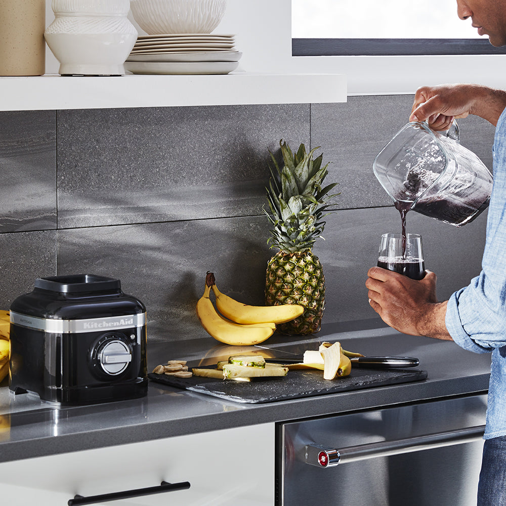 KitchenAid Person pouring red wine into a glass in a kitchen with a pineapple, bananas, and a blender on the counter.