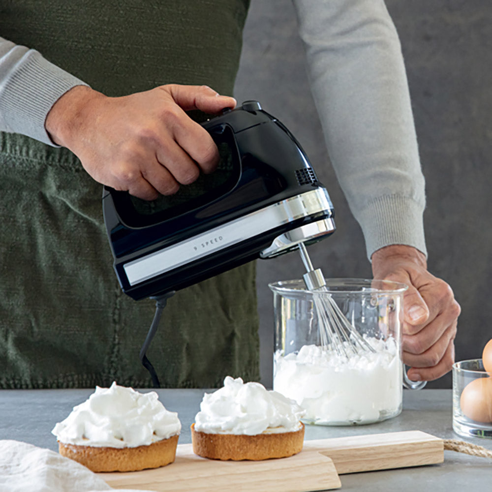 Person using a black hand mixer to whip cream on top of dessert