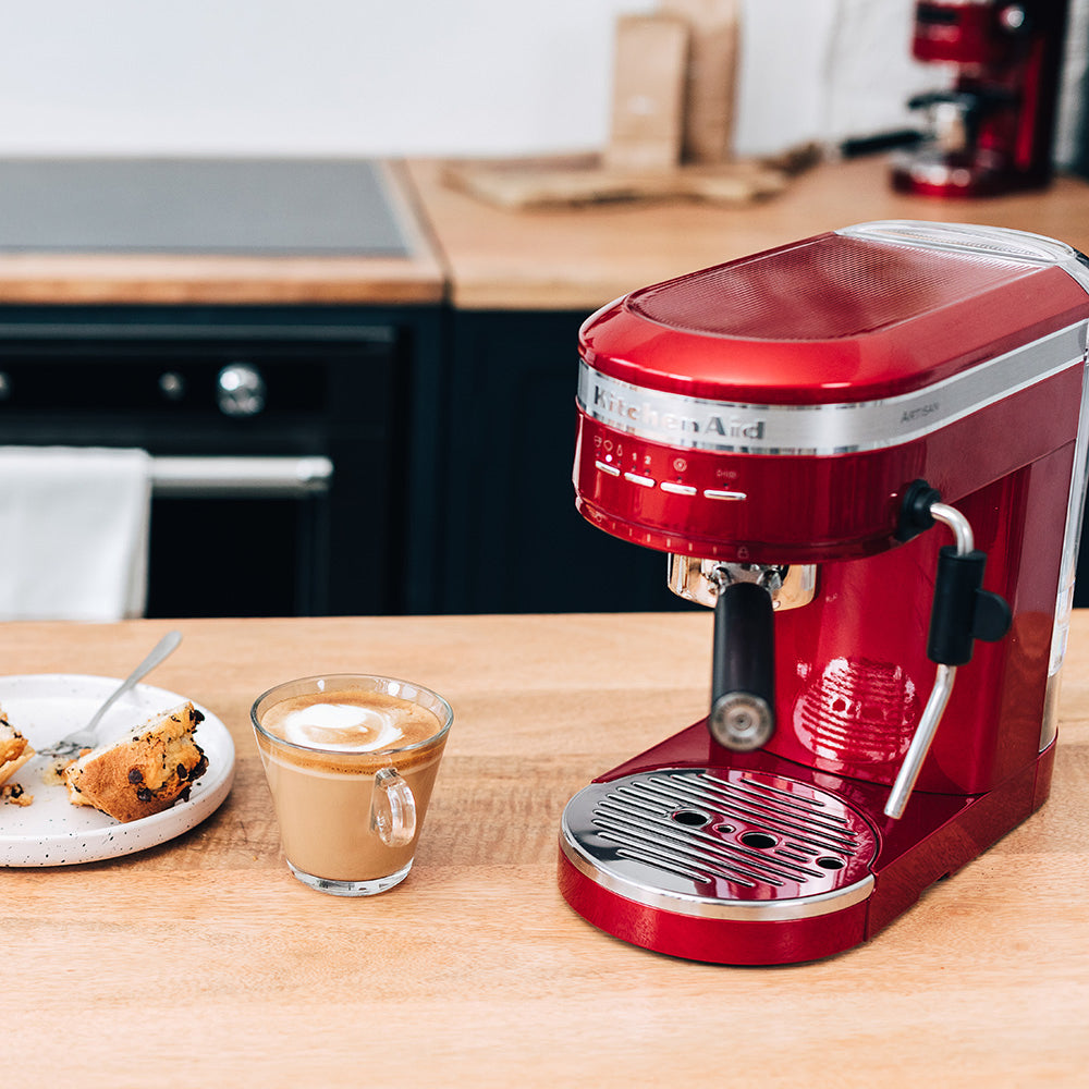 Red KitchenAid espresso machine on a kitchen counter with a glass of coffee and cookies.