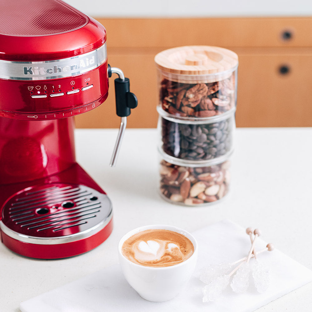 Red coffee machine with a cup of coffee and a container of assorted nuts on a white surface.