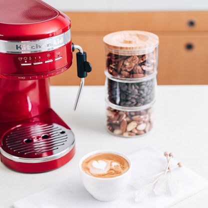 Red coffee machine with a cup of coffee and a container of assorted nuts on a white surface.