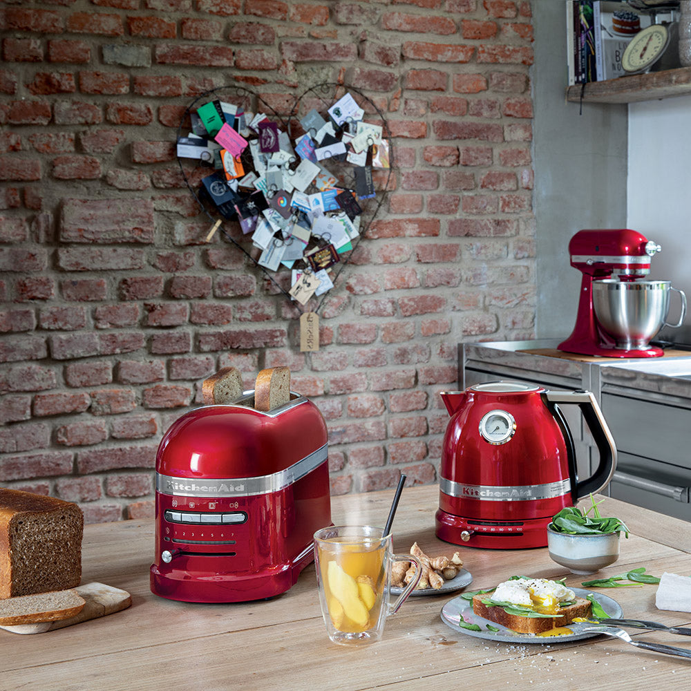 Red kitchen appliances including a toaster and kettle on a wooden table with a brick wall background.