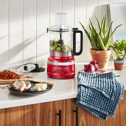Red food processor on a kitchen counter with ingredients and a blue towel.
