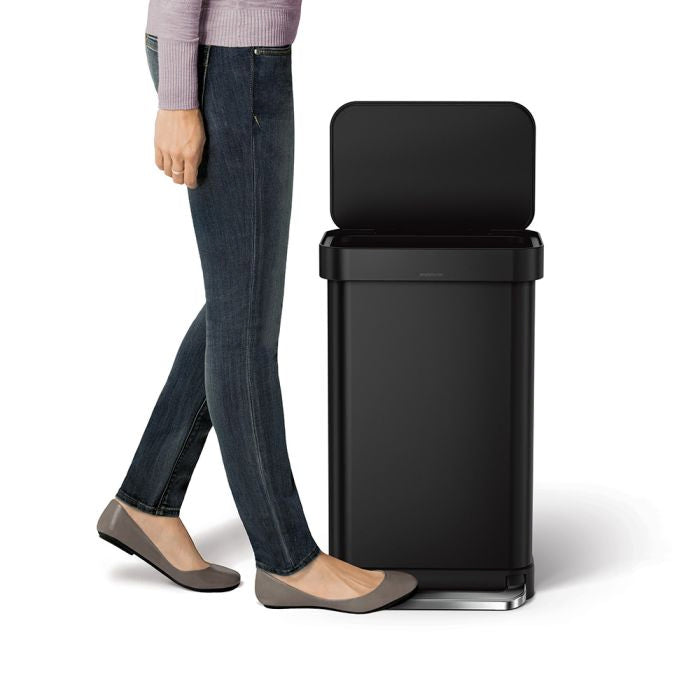Simplehuman Rectangular Pedal Bin with Liner Pocket Black trash bin with a person standing next to it 