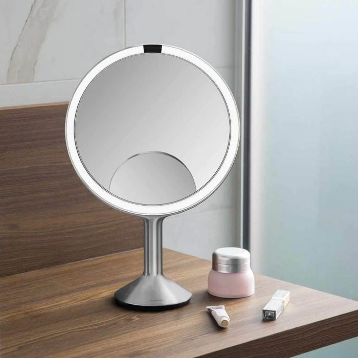 Simplehuman Sensor Mirror Trio Max Round makeup mirror with LED lights on a wooden surface next to skincare products.