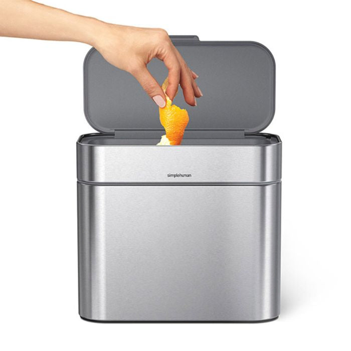 simplehuman Compost Caddy Hand disposing of an orange peel into a silver trash can with 'simplehuman' branding.